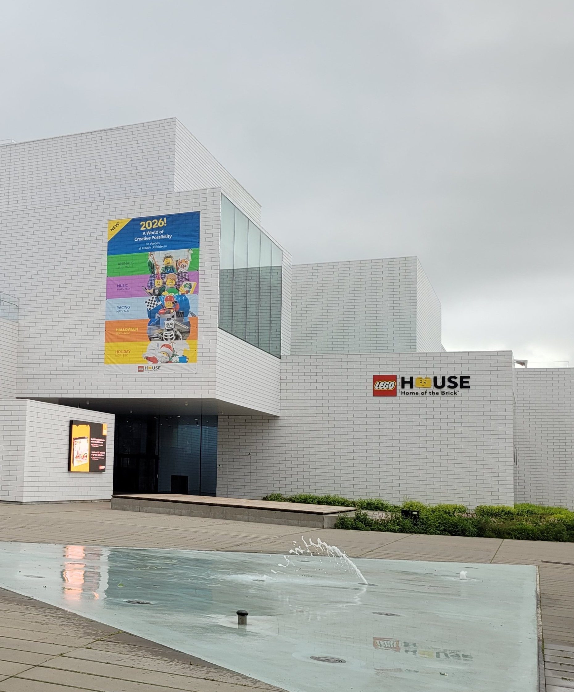 Façade blanche de la LEGO House avec des escaliers et une fontaine à l'avant, des visiteurs se promènent sous un ciel nuageux.